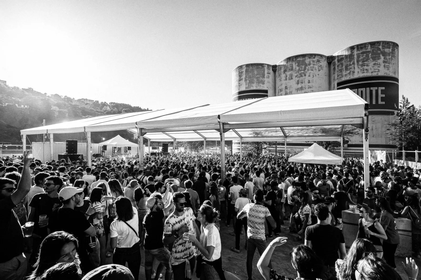 Foule dansant sous une grande tente devant des silos, en noir et blanc